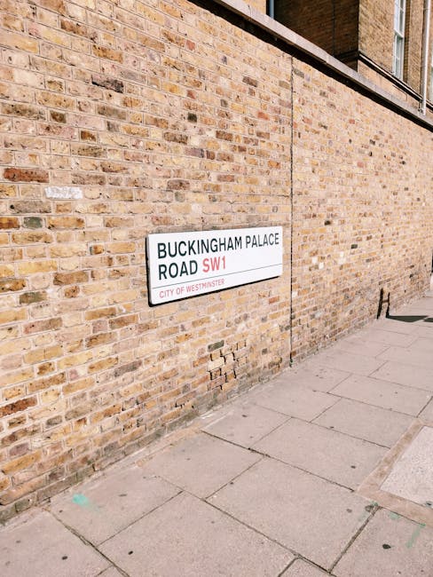 A close-up of a white London street sign for Buckingham Palace Road SW1, mounted on two metal posts, with bold black and red lettering indicating the street name and district. Behind the sign, a refle
